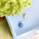 Two small shell-shaped jewelry charms, one light blue and one pale pink, each with a gold-tone loop at the top, displayed on a light blue tray with decorative flowers around it.