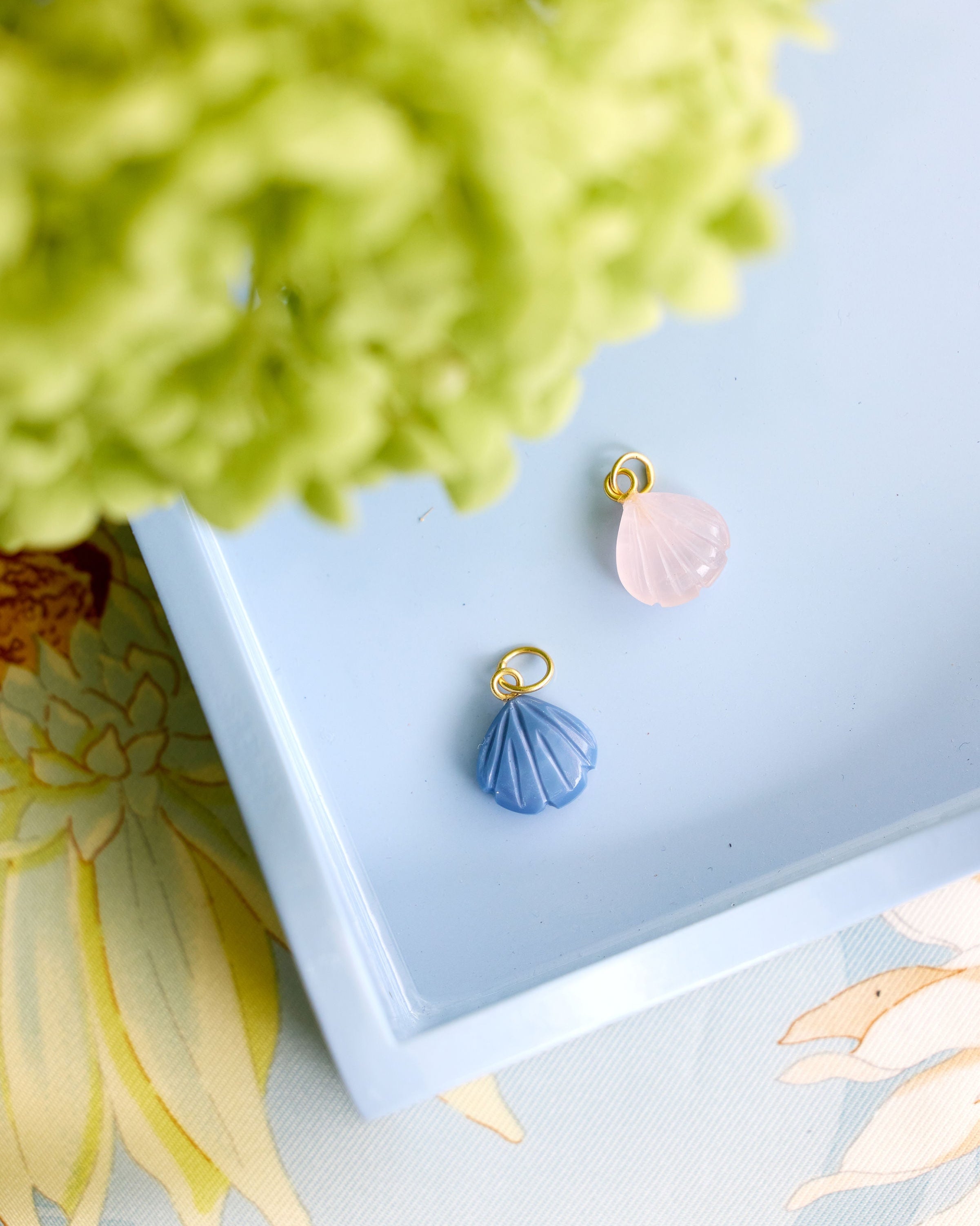 Two small shell-shaped jewelry charms, one light blue and one pale pink, each with a gold-tone loop at the top, displayed on a light blue tray with decorative flowers around it.