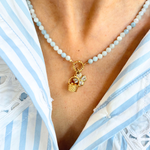 Blue beaded necklace with a golden basket charm and clover charm worn by a person in a white and blue striped garment.
