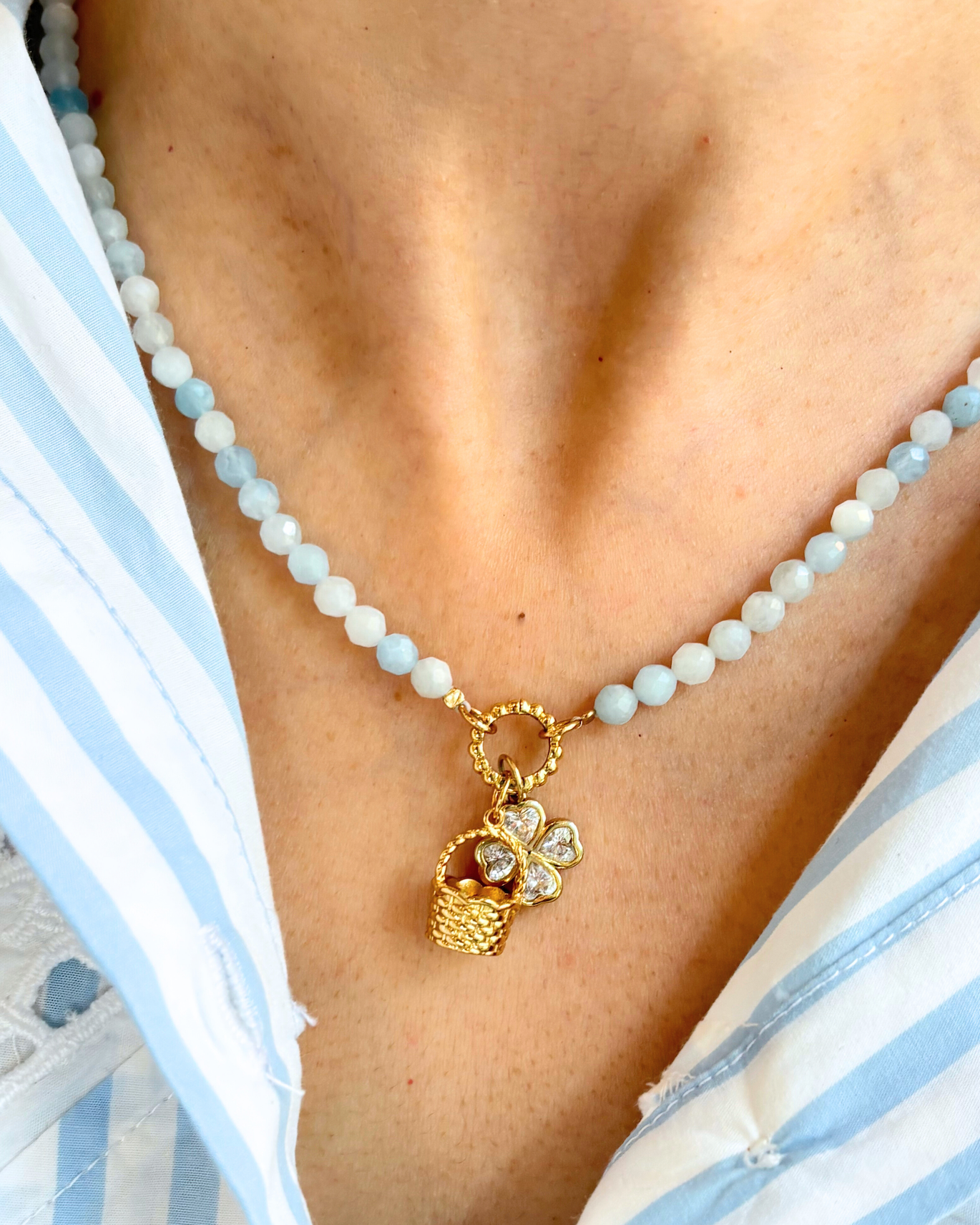 Blue beaded necklace with a golden basket charm and clover charm worn by a person in a white and blue striped garment.