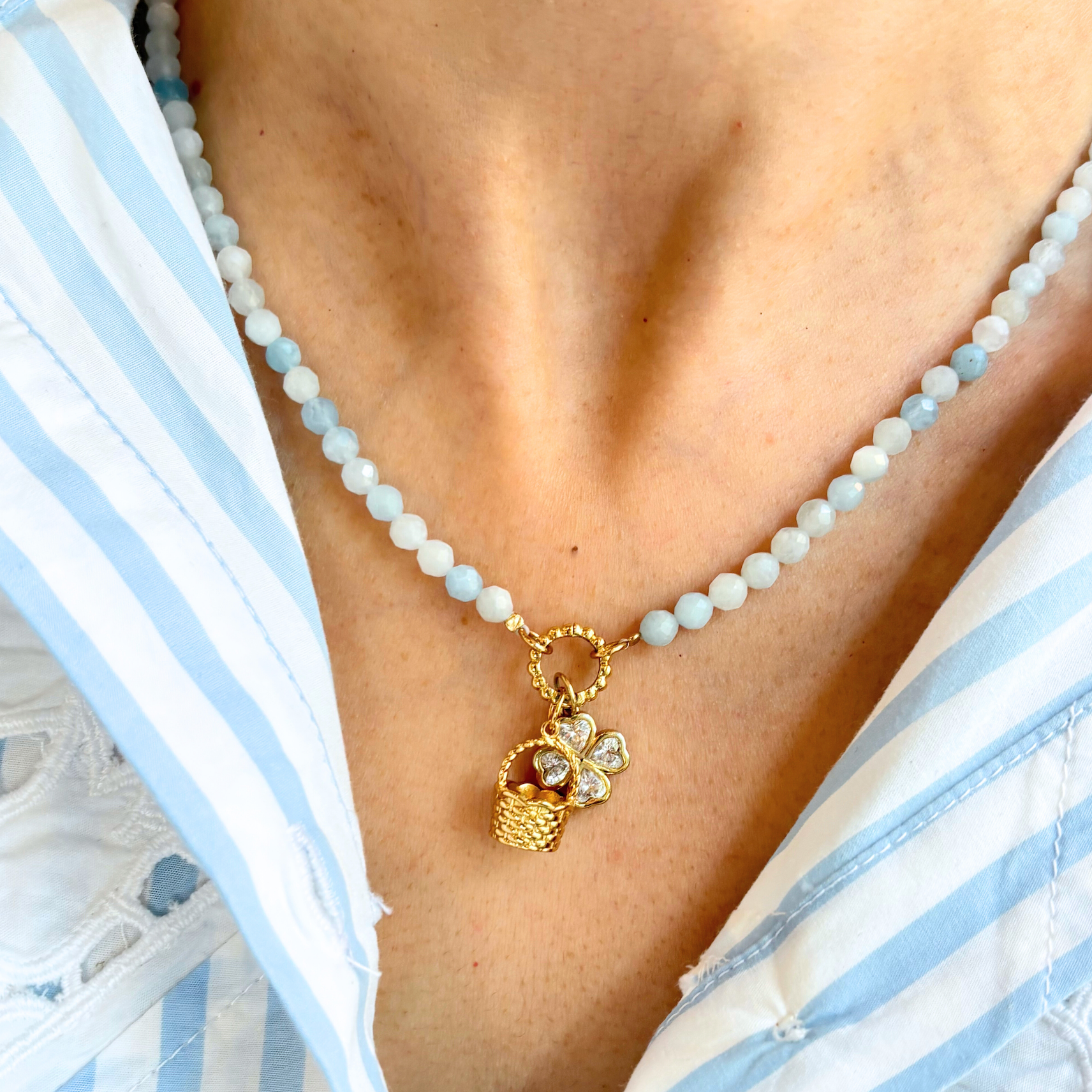 Blue beaded necklace with a golden basket charm and clover charm worn by a person in a white and blue striped garment.