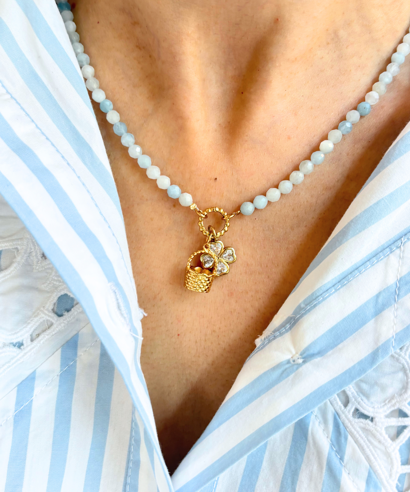 Blue beaded necklace with a golden basket charm and clover charm worn by a person in a white and blue striped garment.