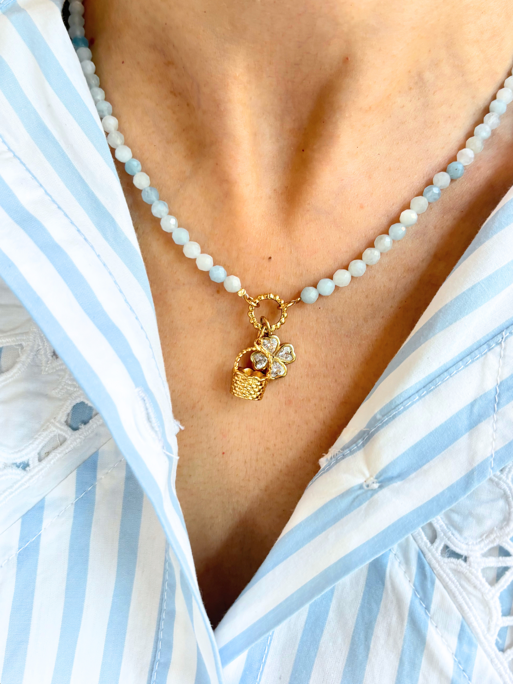 Blue beaded necklace with a golden basket charm and clover charm worn by a person in a white and blue striped garment.