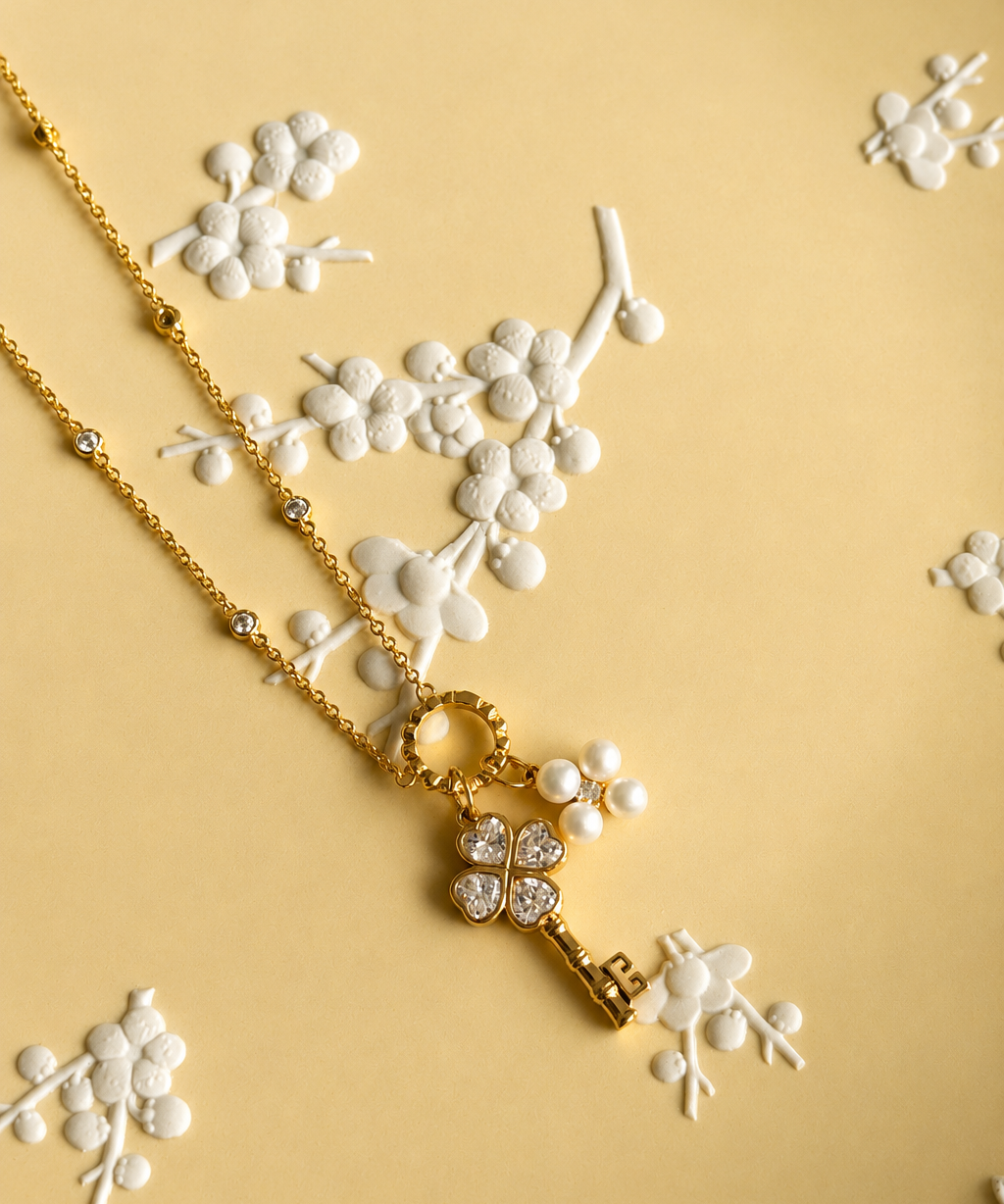 A gold key pendant and pearl clover necklace with clear rhinestones, displayed on a textured yellow background decorated with white 3D floral elements.