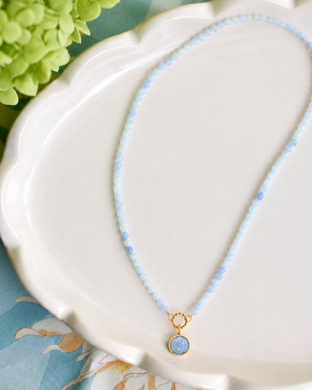 A delicate beaded necklace with light blue beads and a gold-tone pendant featuring a round light blue stone, displayed on a white scalloped dish.