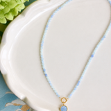 A delicate beaded necklace with light blue beads and a gold-tone pendant featuring a round light blue stone, displayed on a white scalloped dish.
