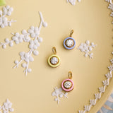 Three small circular pendant charms with gold-tone rings and clear stones, each set in a different pastel color (blue, yellow, pink), arranged on a pale yellow decorative plate with white floral relief details.