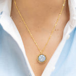 Gold necklace with a circular blue pendant worn by a person in a light blue shirt.