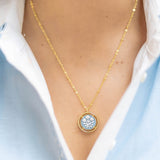Gold necklace with a circular blue pendant worn by a person in a light blue shirt.