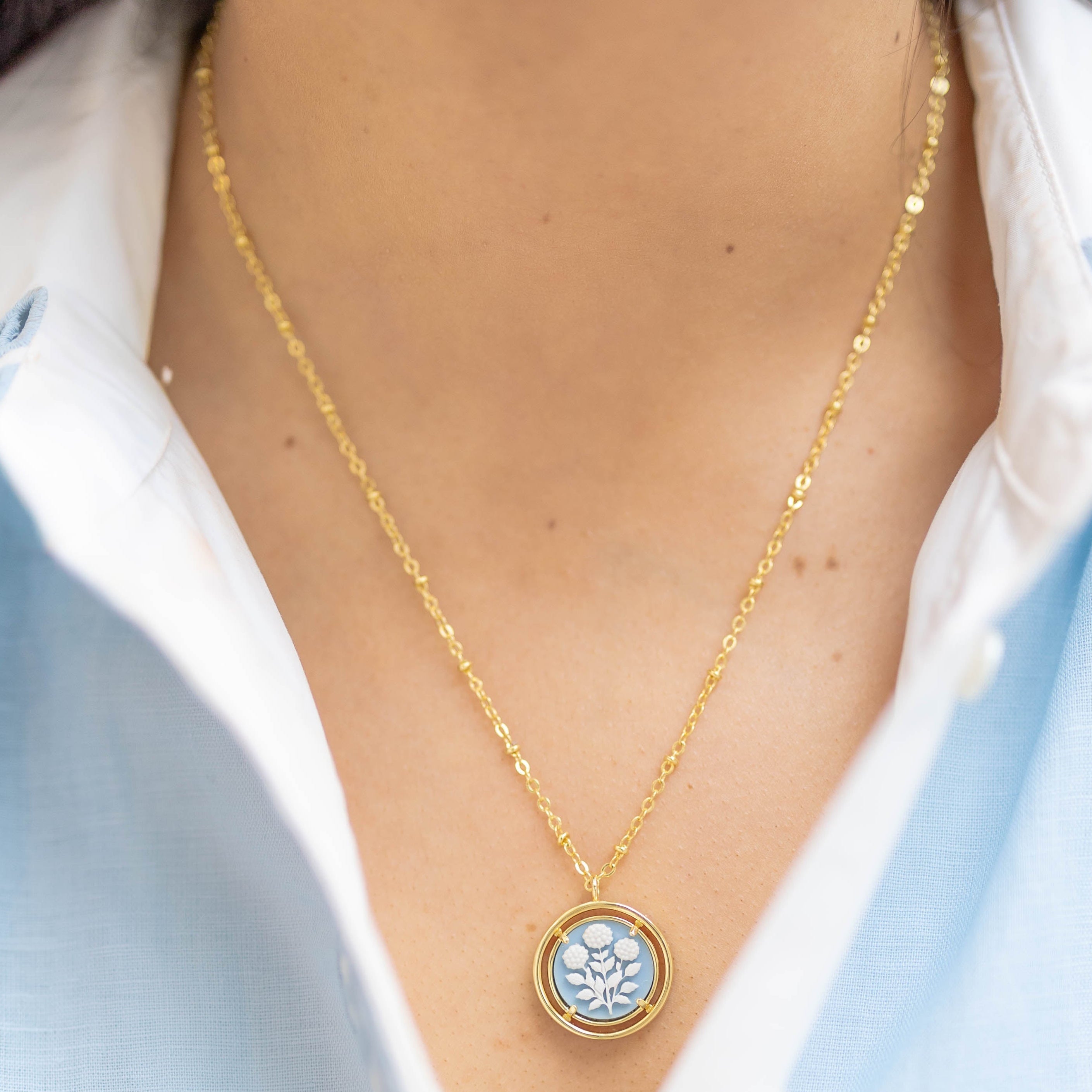 Gold necklace with a circular blue pendant worn by a person in a light blue shirt.