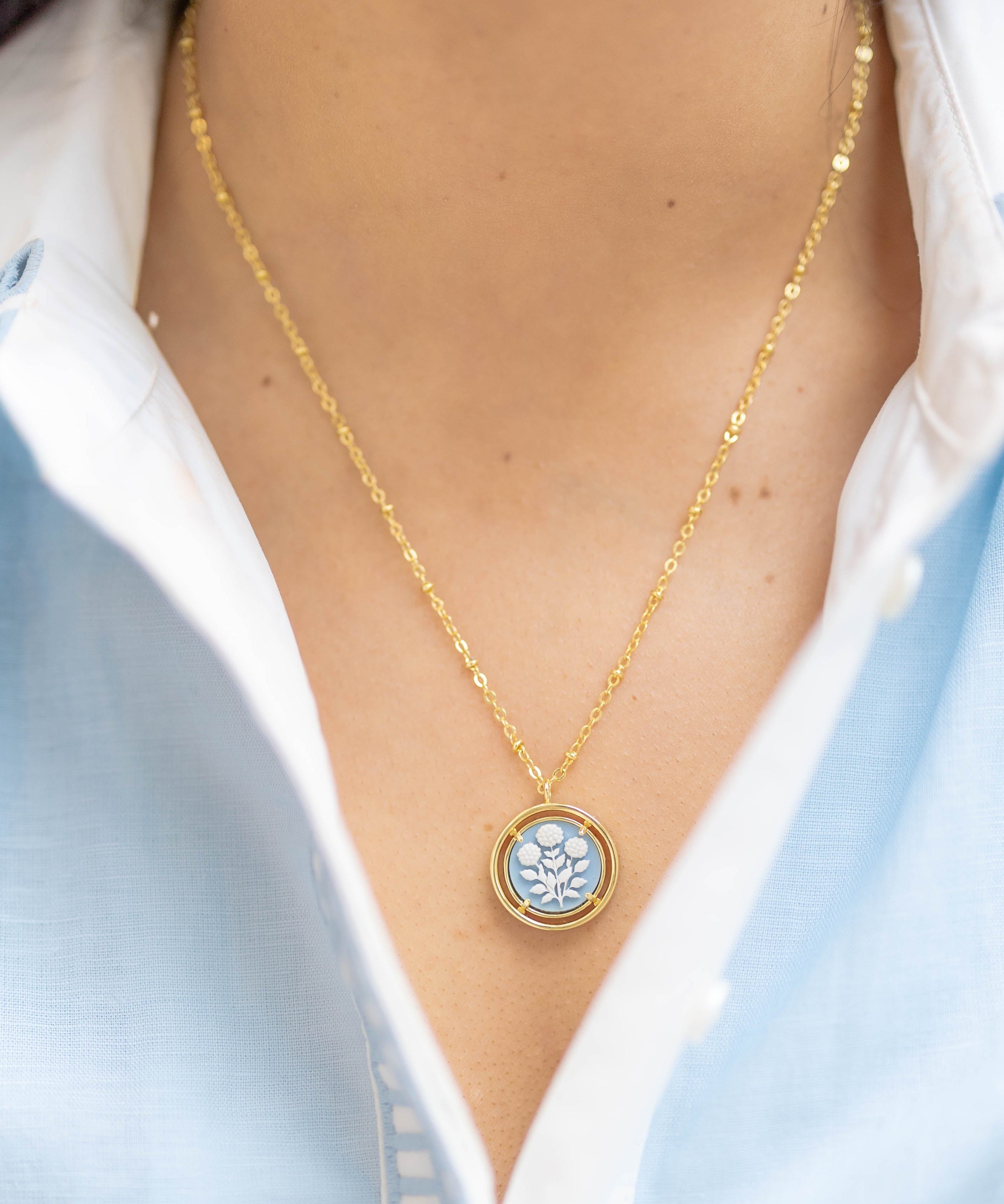 Gold necklace with a circular blue pendant worn by a person in a light blue shirt.