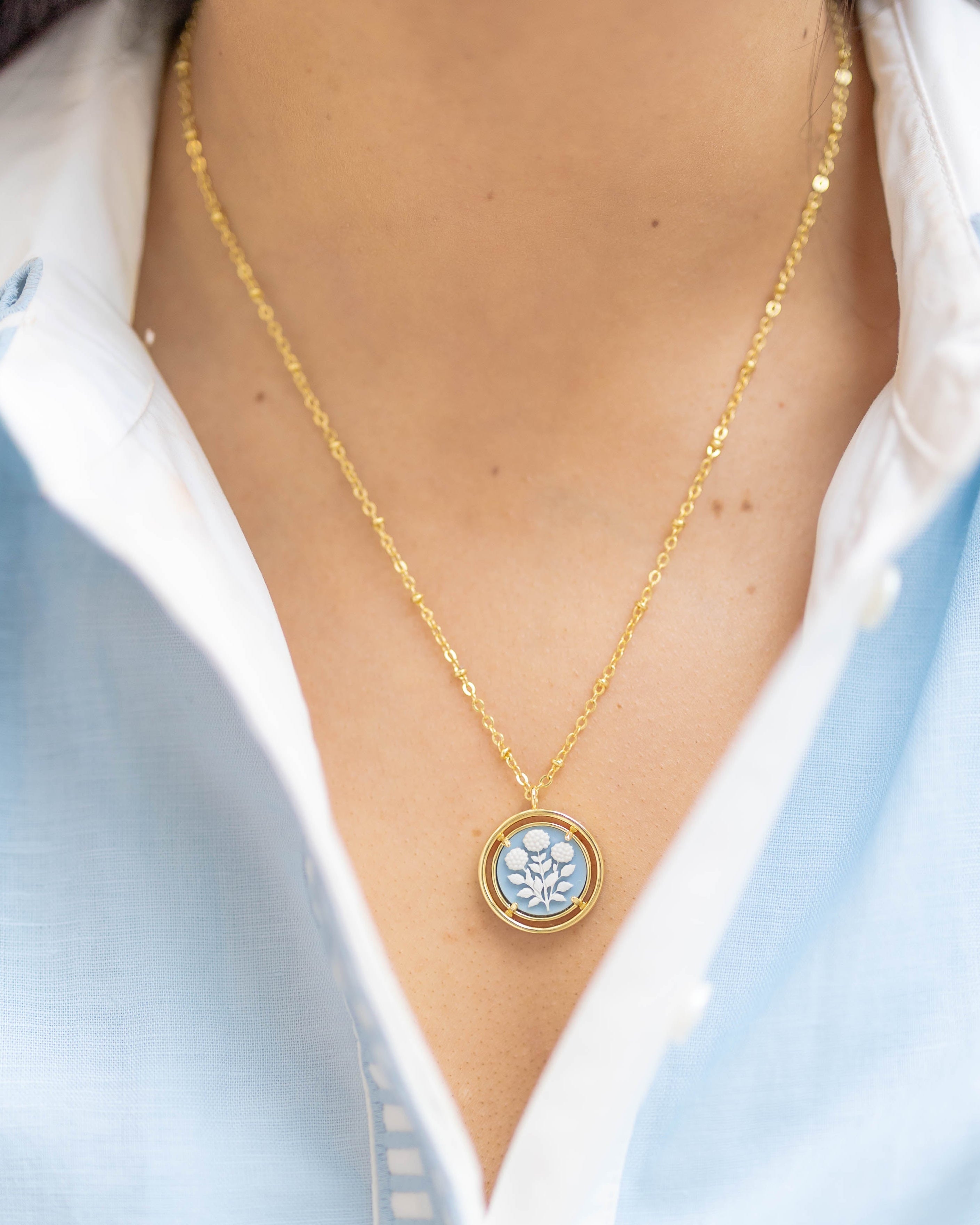 Gold necklace with a circular blue pendant worn by a person in a light blue shirt.