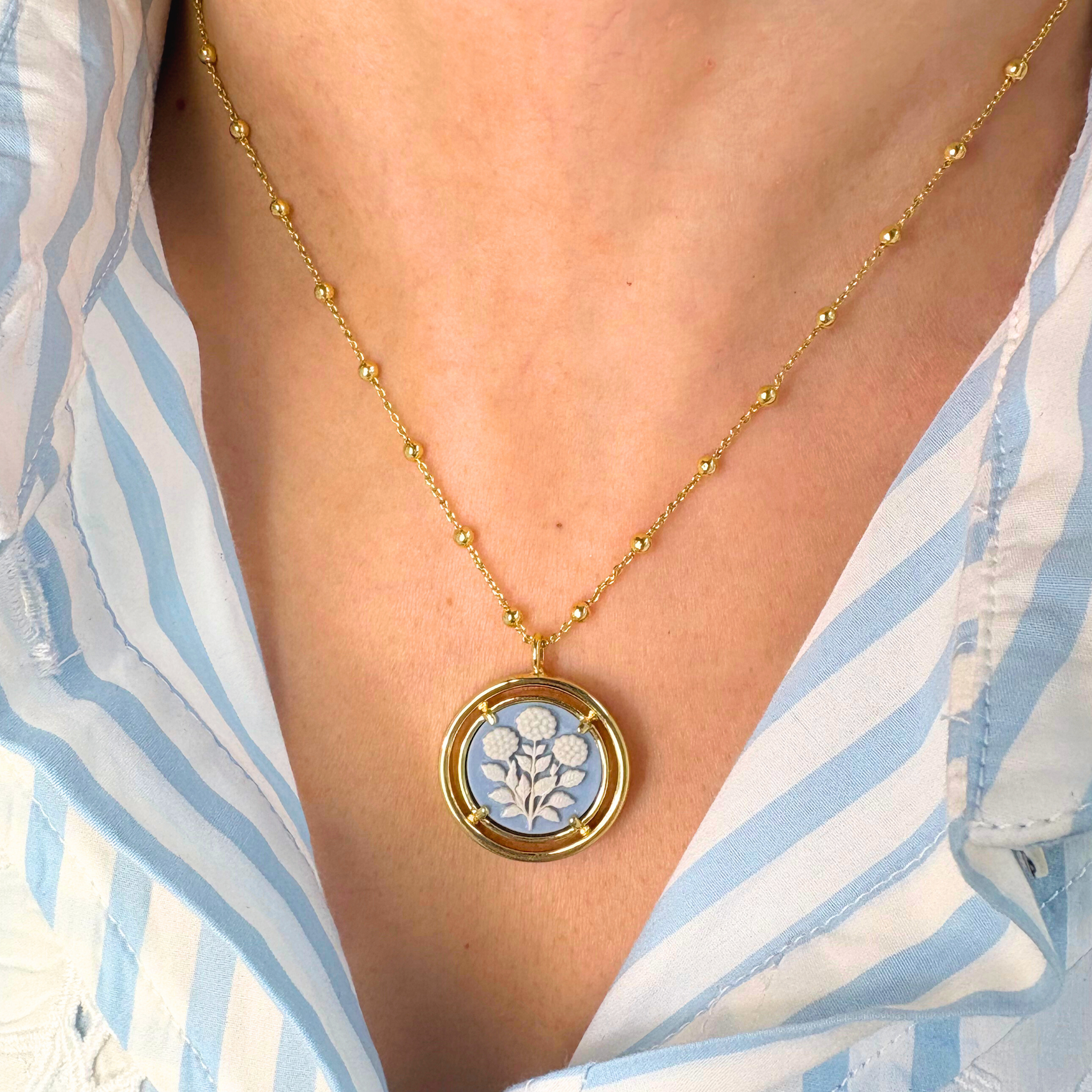 Blue cameo pendant necklace worn by a person in a blue and white striped shirt.