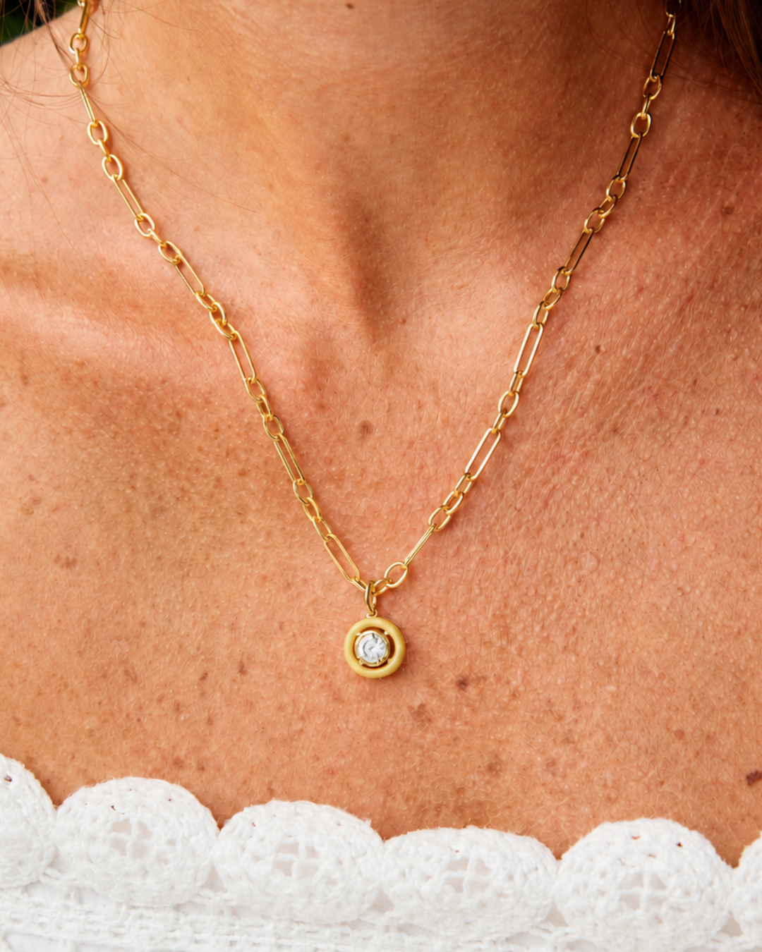 A close-up of a gold chain necklace with a small round bezel-set clear gemstone pendant, worn on a model's neck above a white lace top.