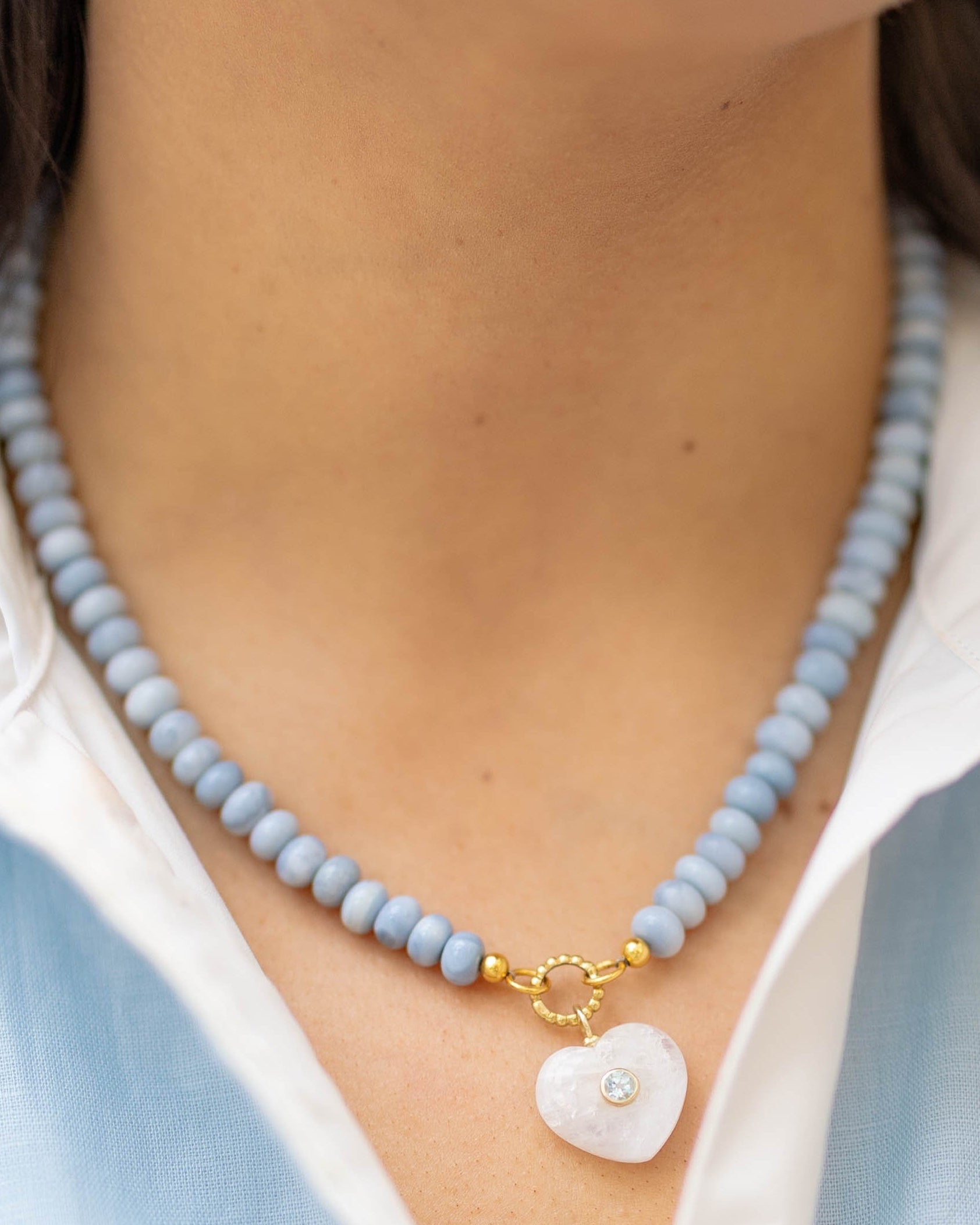 moonstone heart charm on a blue beaded necklace on a woman wearing a blue and white shirt