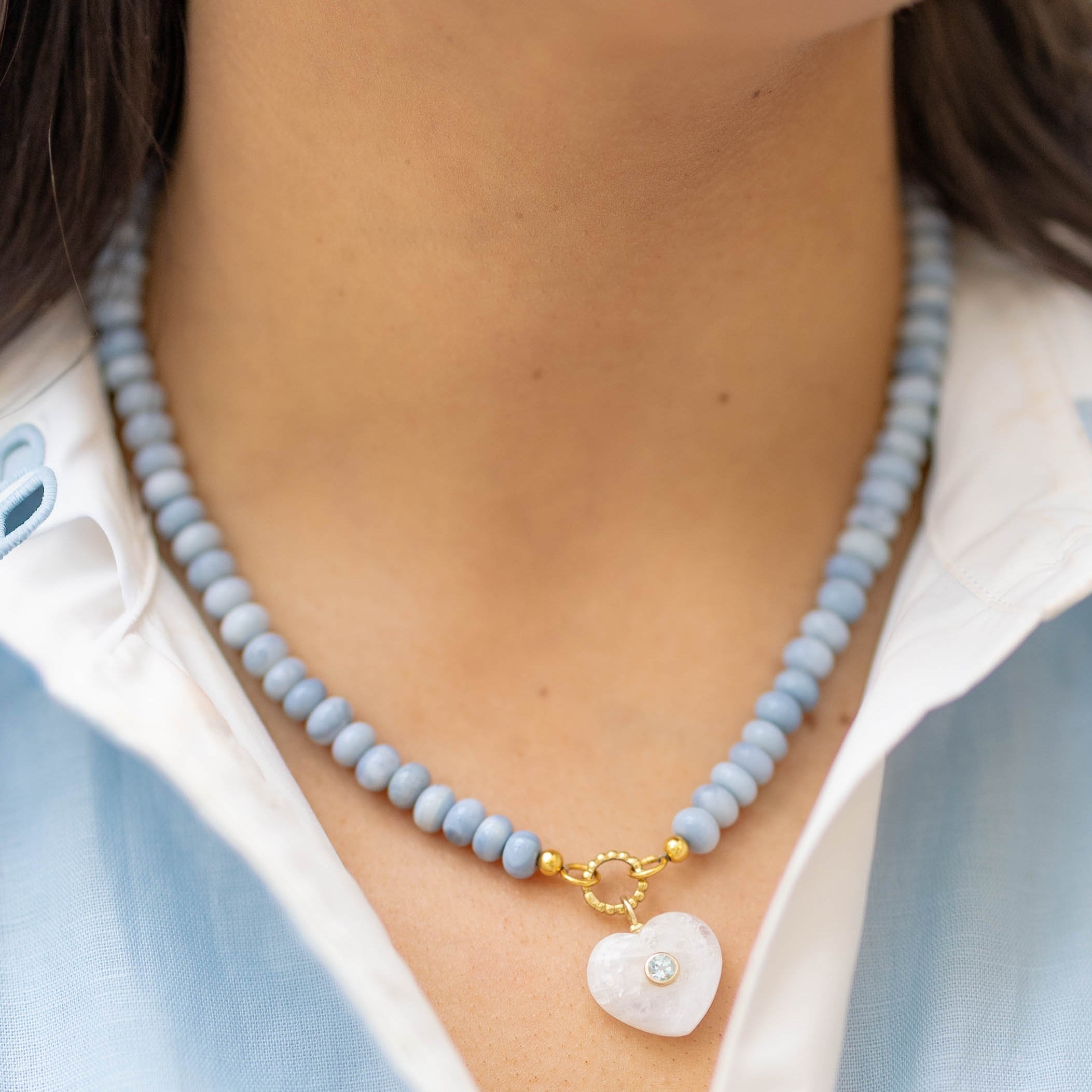 moonstone heart charm on a blue beaded necklace on a woman wearing a blue and white shirt
