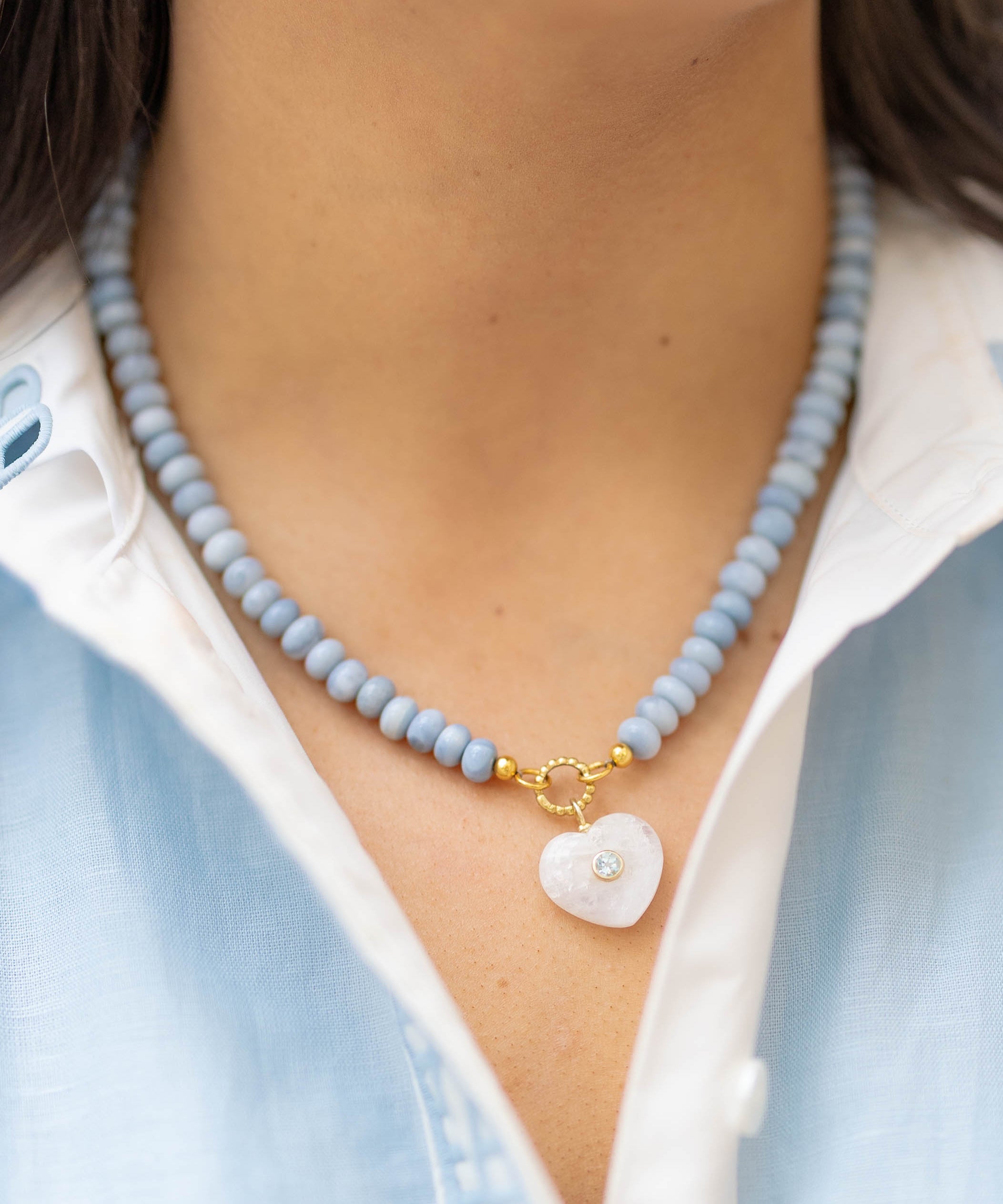 moonstone heart charm on a blue beaded necklace on a woman wearing a blue and white shirt