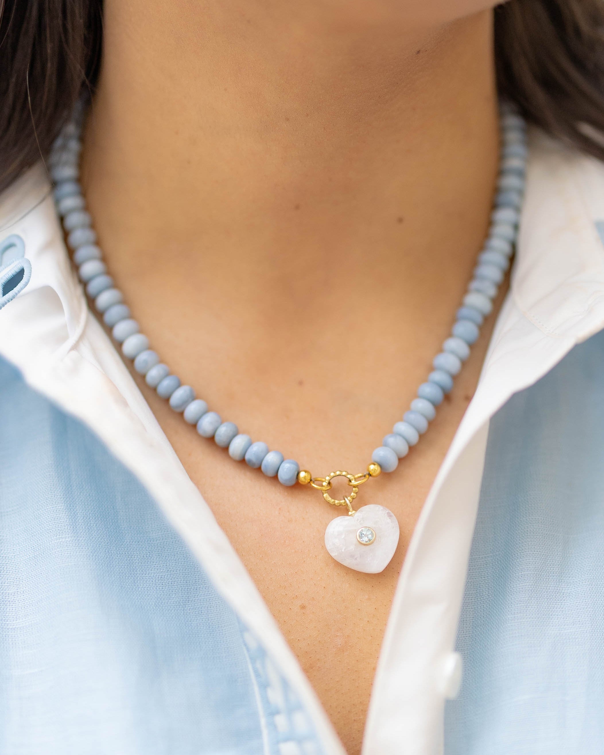 moonstone heart charm on a blue beaded necklace on a woman wearing a blue and white shirt