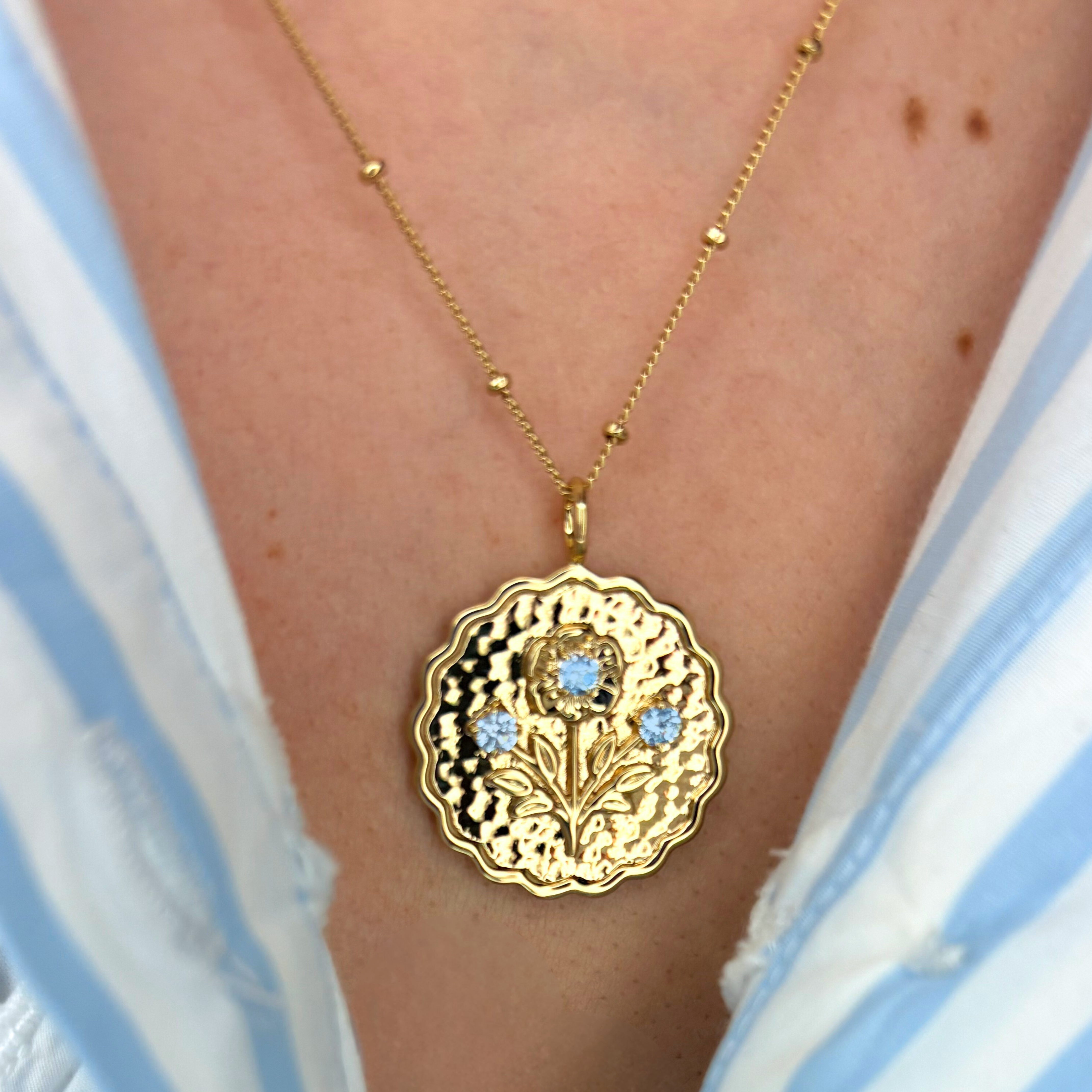 Gold necklace with a decorative pendant featuring blue stones, worn by a person in a striped garment.
