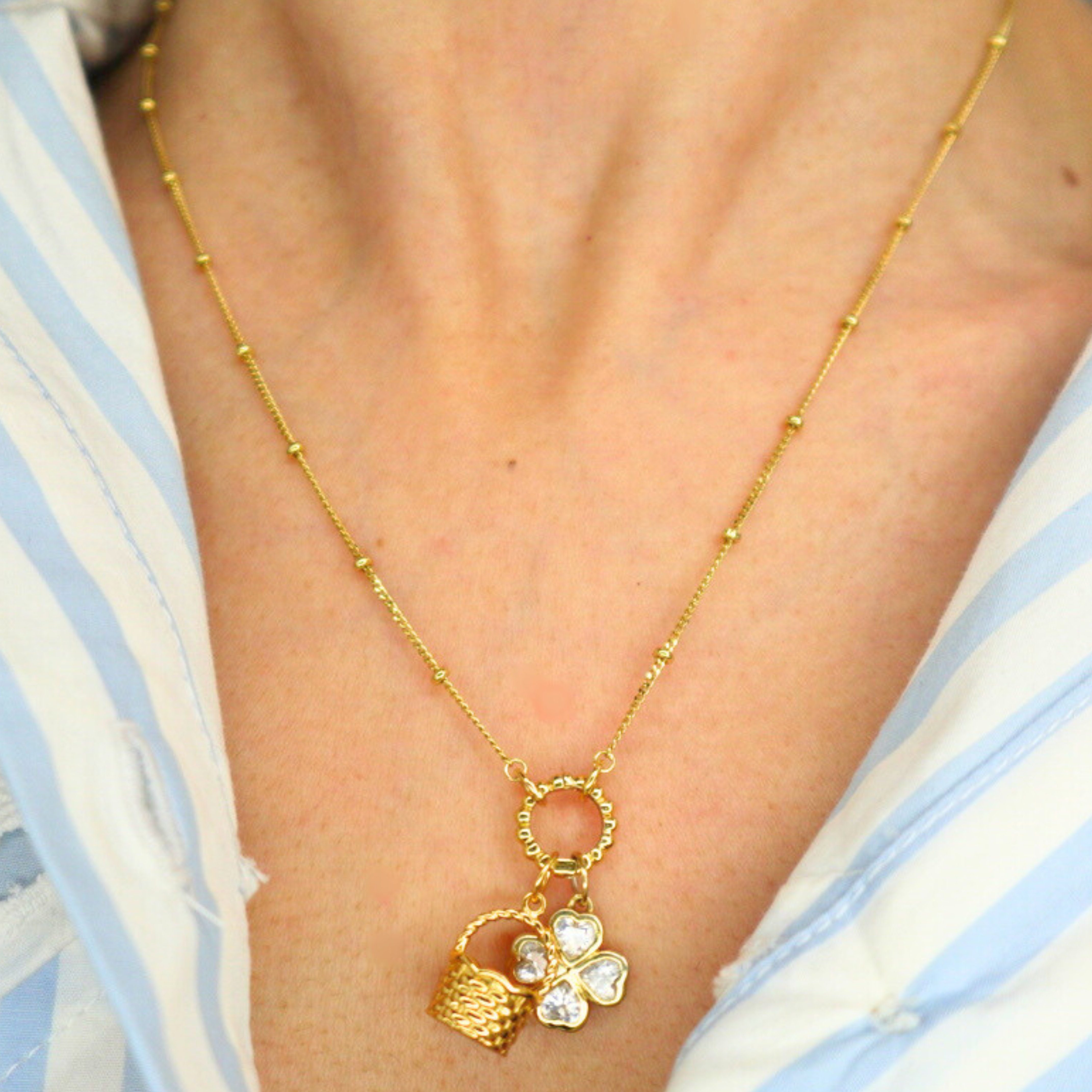 golden necklace with a basket charm and clover charm on a person wearing a blue and white striped shirt