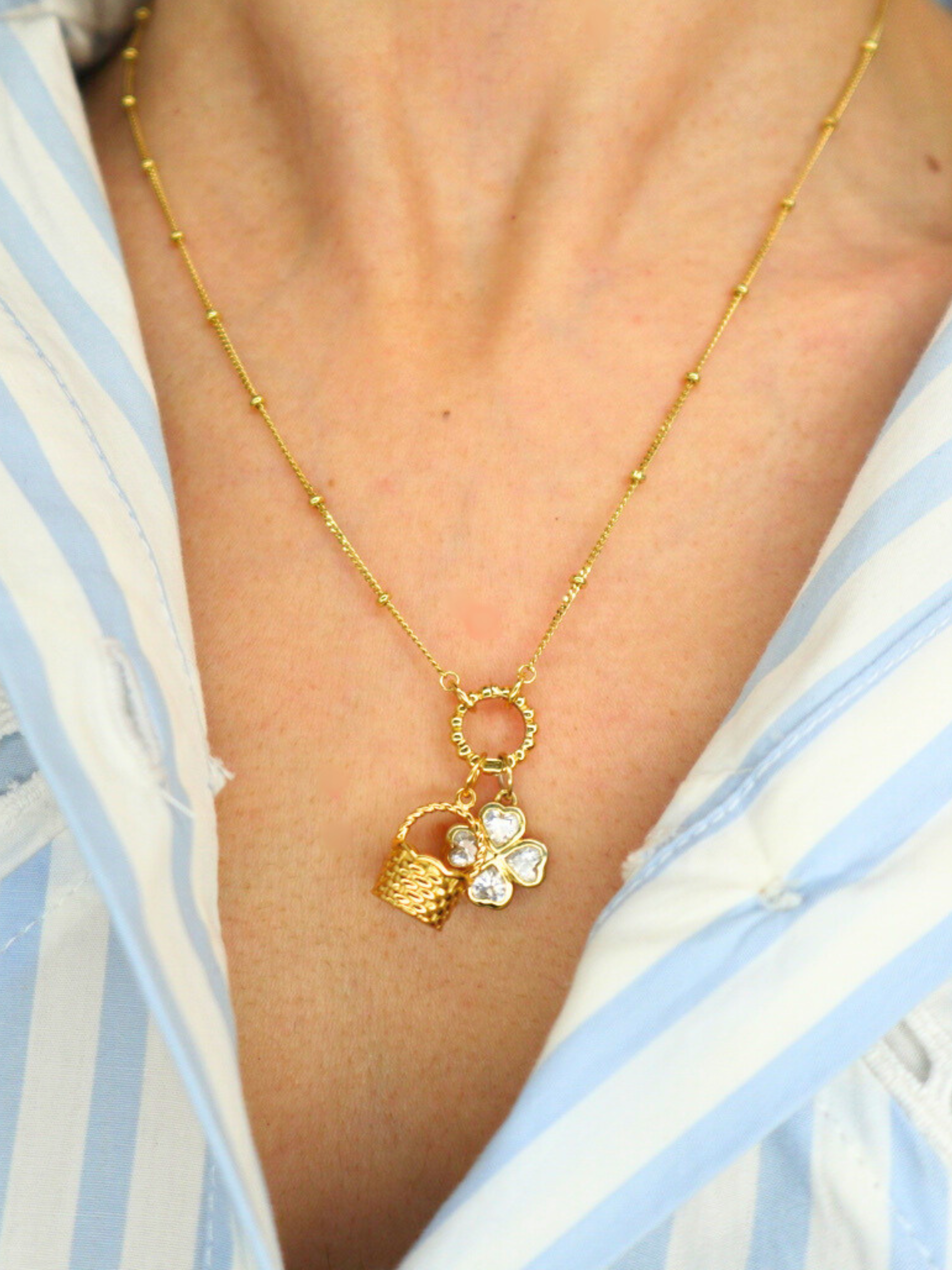 golden necklace with a basket charm and clover charm on a person wearing a blue and white striped shirt