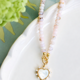 A beaded necklace with pale pink and white marbled beads and a gold heart-shaped pendant with a mother-of-pearl center, displayed on a white ceramic dish.