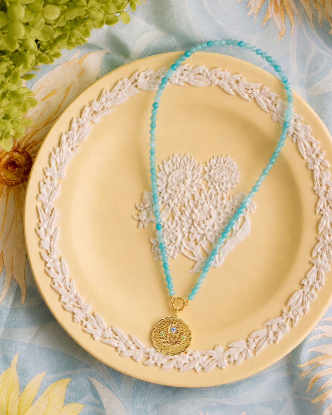 A turquoise beaded necklace with a gold circular pendant lies on a pale yellow plate with white floral detailing, styled with dried flowers and a light blue fabric background.