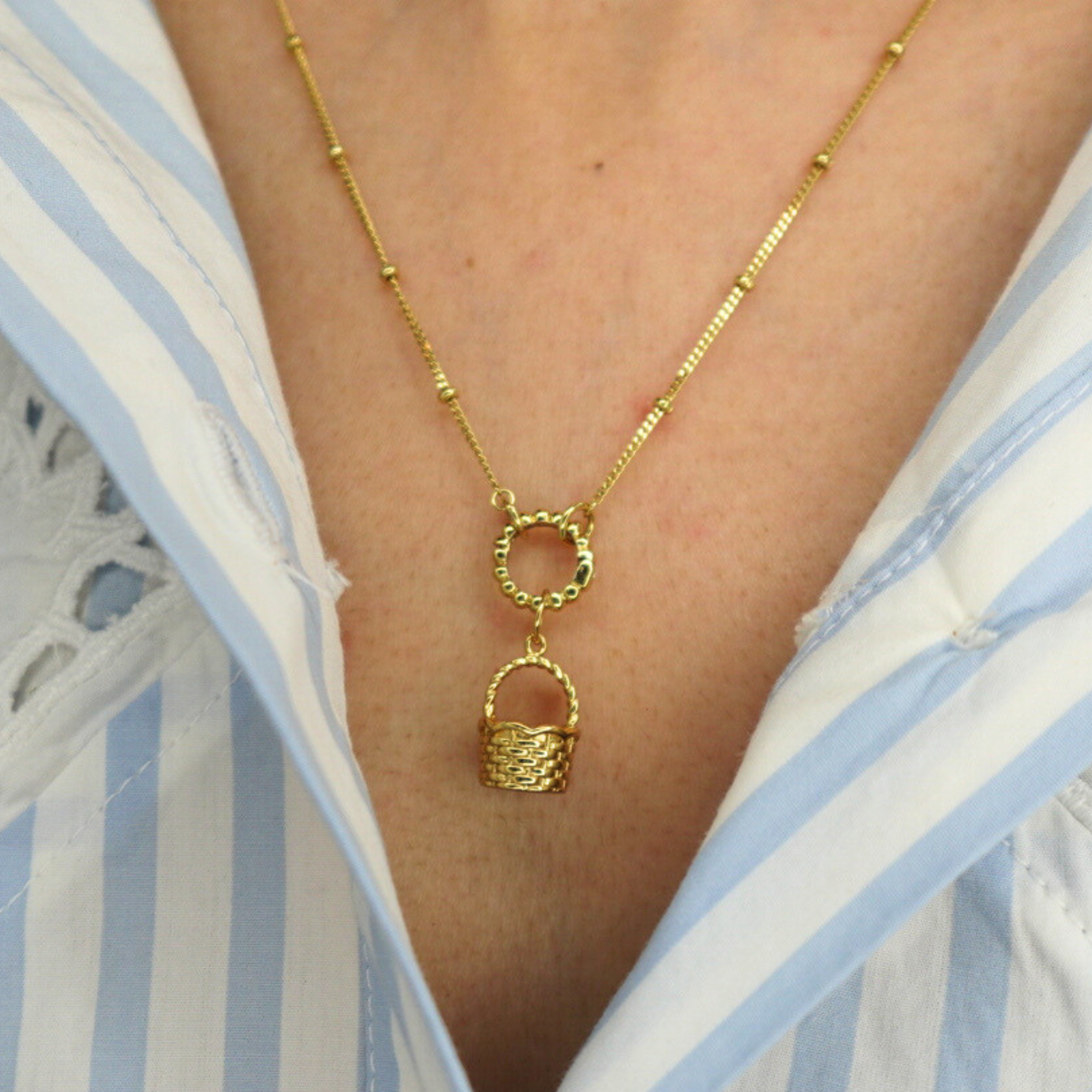 golden basket charm on a golden necklace worn by a person in a blue and white striped shirt