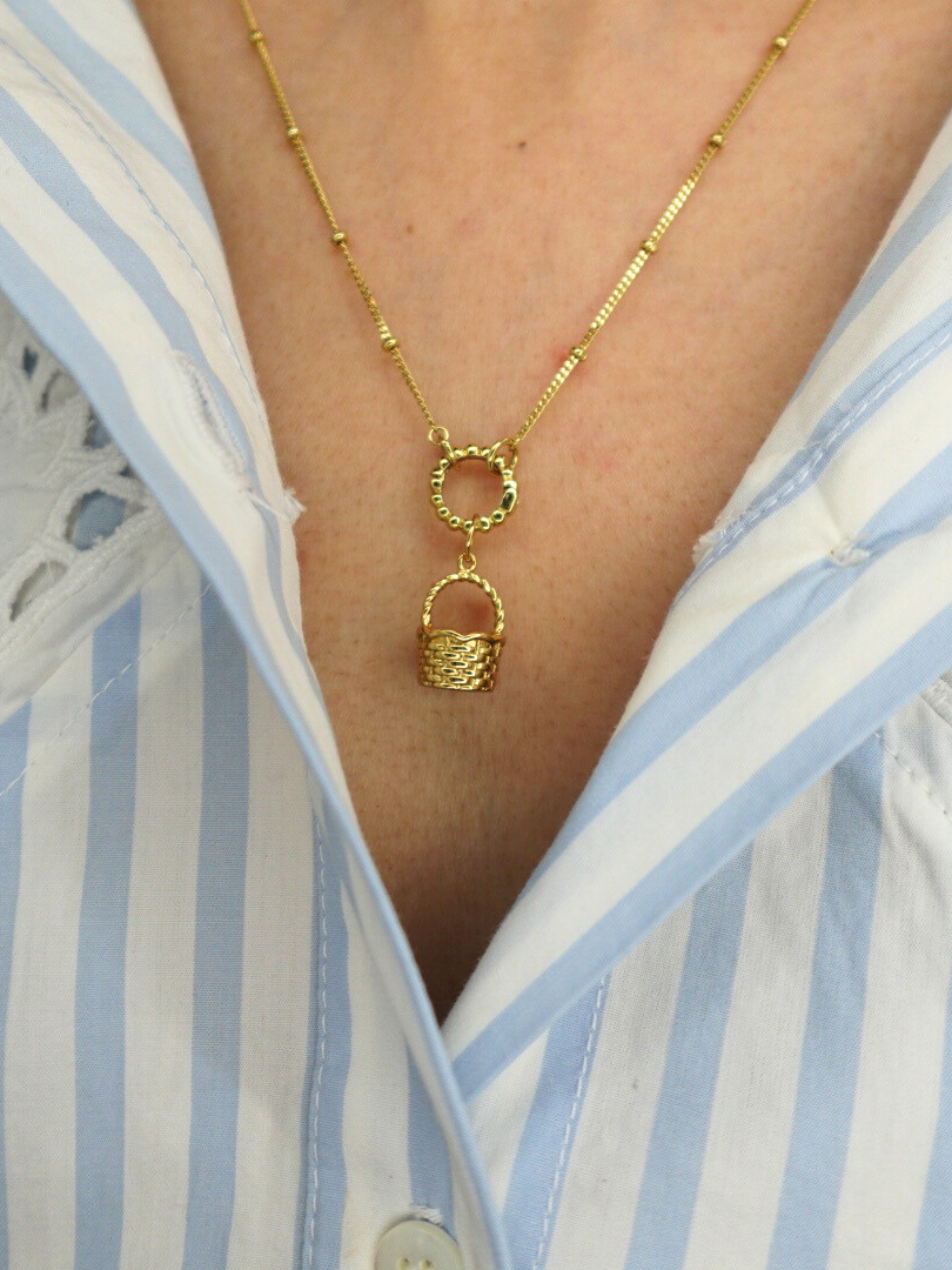 golden basket charm on a golden necklace worn by a person in a blue and white striped shirt